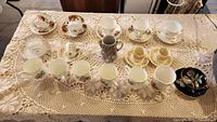 Full view of 18 cups, saucers, and mugs arranged on lace tablecloth, showing variety in shapes, sizes, and floral patterns