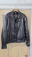 Front view of black leather jacket hanging on wooden hanger against door.