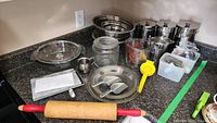Overview of vintage kitchenware lot showing bowls, utensils, rolling pin, measuring cup, and glass containers on countertop.
