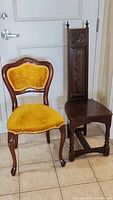 Pair of antique chairs. Left chair is upholstered in yellow velvet with carved wooden frame. Right chair is tall wooden with carved floral panels on backrest.