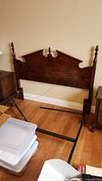 Full view of queen size wooden headboard attached to metal bed frame with parts detached and resting against side wall. Wood is dark stained with three finials.