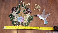 Photo showing both stained glass suncatchers laid on a wooden table with a tape measure for size reference. Floral pineapple design approximately 8 inches wide, detailed stained glass work with red roses and green leaves, and a hummingbird figure with pastel colored glass.