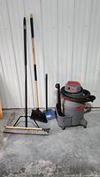 Full view showing the vacuum, broom, dustpan with brush, and floor squeegee against a white wall.