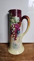 Tall handpainted tankard pitcher with grapevine design, showing front side, highlighting grapes and handle.