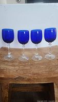 Photo showing four cobalt blue goblets with clear faceted stems and clear round bases standing on a wooden surface.