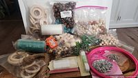 Wide view of crafting supplies showing multiple bags of various sized wooden beads, multiple bags and packages of wooden rings, flat waxed thread spools in green and white, green glass flower beads, a packet of safety pins, and natural fiber string bundles.