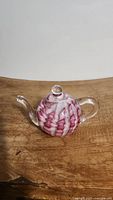 Front-left angle view of a glass teapot paperweight with pink and white swirls on a wooden surface.