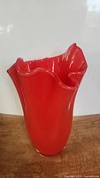 Red handmade glass vase with wavy top edge, smooth finish, standing on wooden surface.