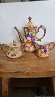 Front view of the Lefton China hand-painted tea set showing teapot, sugar bowl, and creamer with detailed fruit and gold designs