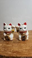 Front view of two white ceramic Maneki Neko lucky cat figurines with red and gold collars, each holding a gold coin with black characters. Left cat paw raised right, right cat paw raised left.