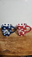 Front view of the blue Mickey Mouse and red Minnie Mouse polka dot coffee mugs on a wooden surface.