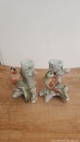 Pair of vintage hand-painted ceramic bird candle holders on wooden surface, front view showing details of birds and tree stump bases.