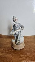Side view of vintage porcelain clown figurine music box showing the clown playing accordion, gray and white colors, wooden base.