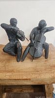 Two dark-toned vintage sculptures of seated musicians, one playing violin and one cello, on wooden surface, showing full figures and detailed forms.