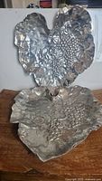 Two vintage aluminum grape leaf shaped trays with grape cluster embossing, one standing vertically and one laying flat on wood surface.