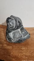 Front and side angled view of the dark gray soapstone Inuit sculpture showing the figure's face, hooded parka, arms, and detailed carved textures.