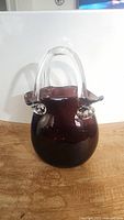 Front angled view of the purple/violet hand blown glass purse vase, showing the glossy rounded body and clear handle.