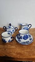 Photo showing two teacups with saucers, sugar bowl with lid, and creamer, all decorated in blue floral flow blue pattern on cream ironstone porcelain.