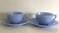 Two blue Pyrex Delphine cups with matching saucers shown side by side on a neutral surface.