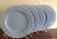 Five blue Pyrex Blue Delphine dinner plates arranged standing on clear plastic plate holders, side view showing fluted pie crust edges and consistent light blue color.