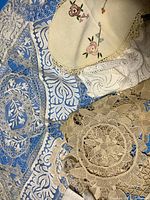 Various hand made lace pieces spread out on a surface showing details of pattern, texture, and variety of lace styles including a beige crocheted doily and embroidered linen doily.