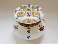 Front view of round white milk glass dresser trinket box with hand-painted colorful floral and gold decoration, hinged brass collar visible around lid.