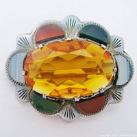 Close-up image of antique Scottish brooch showing faceted orange paste centerpiece with engraved silver surround and multicolor inset stones, front view.