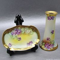 Two decorative vanity items with purple violet floral designs and gold rims. Left item is an oval pin dish on a black stand; right item is a tall conical hat pin holder.