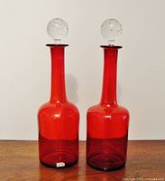 Pair of bright cherry red glass decanters with clear bubble glass stoppers shown upright on wooden surface against white background.
