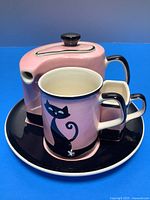 Pink tea set components (teapot, cup, creamer) arranged on round plate, showing black cat design and glossy finish