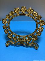 Full front view of the oval gold coloured metal frame showing floral decoration around the border and curled metal base.