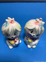Two anthropomorphic ceramic salt and pepper shakers shaped like Maltese puppy dogs with pink bows and flowers in hair, viewed from the front on blue background.
