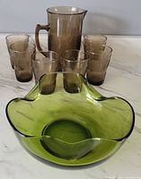 Green glass chip and dip bowl in front of six smoky grey glasses and a pitcher on marble countertop