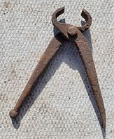 Antique farrier pliers shown resting on a textured surface, highlighting the full open jaws and overall rusted condition.
