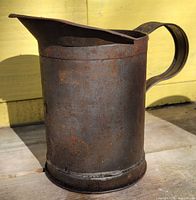 Side view of the cylindrical tin measuring cup showing heavy rust and patina, with a spout and large handle.
