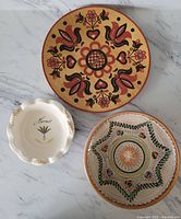 Front view of three decorative plates: terra cotta plate with floral motif; ceramic plate with intricate design; small cream colored ceramic plate with 'Hernes' and a leaf design.