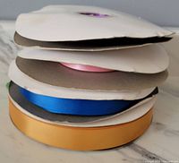 Stack of four rolls of crafting ribbon in coral pink, gold, purple, and blue with cardboard holders.
