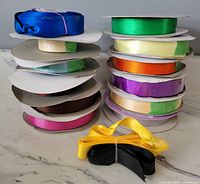 Stack of satin ribbons in multiple colors including green, blue, yellow, orange, purple, cream, brown, pink and black, all 7/8 inch wide with mostly full and some open rolls.