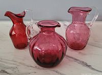 Three small cranberry glass pitchers placed side by side on a white marble surface against a gray background. The pitchers vary in shape, with one round, one narrow-neck with handle, and one taller with spout. The glass varies from deep red to pink tones, all translucent.