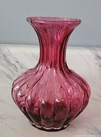 Cranberry-colored ribbed glass vase with bulbous shape and flared neck shown on white marble surface against plain background.