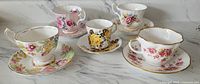 Five floral-themed English bone china teacups with matching saucers set on marble surface showing front view.