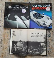 Photo shows three hardcover car books laid out on a carpeted floor. Covers of "Ultimate Autos" and "Ultra Cool Ride" visible, plus an open page from "Vintage Cars Motoring in the 1920s" showing a black and white photo of vintage cars.
