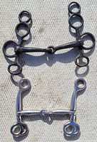 Two Pelham horse bits on a woven surface, one darkened metal and one polished silver, both featuring jointed mouthpieces and multiple rings on shanks.