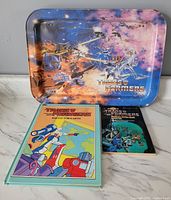 Original 1984 metal Transformers TV tray featuring colorful battle artwork with Optimus Prime and other characters, placed behind two vintage Transformers books on a white surface.