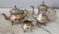 Silver plate tea pot, coffee pot, and creamer with ornate finials and three-legged feet placed on marble surface.