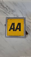 Front view of the square vintage AA car badge with yellow enamel and bold black 'AA' letters, showing some surface marks.