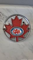Front view of the circular metal grill badge showing the red maple leaf and CAA logo in the center on a marble surface.