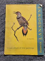 Yellow cover album titled 'Fenwick Lansdowne - A new album of bird paintings' featuring an illustration of a bird (Red-Shafted Flicker) perched on a branch.