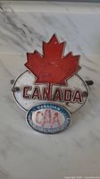 Front view of the vintage metal CAA car badge showing red maple leaf, CANADA text, and circular CAA emblem.