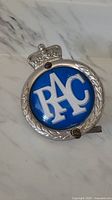 Front view showing blue circular badge with white 'RAC' lettering, silver edging, and crown emblem on top.